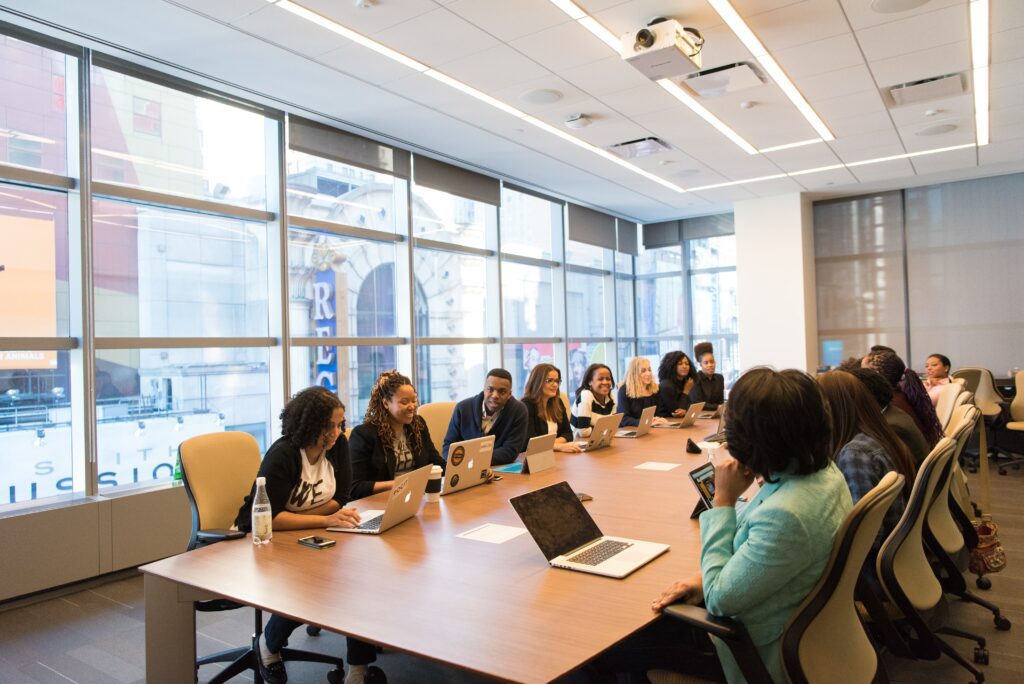 Photograph of diverse group of smiling and talking people with laptops sitting at a long table in a conference room with lots of natural window light.